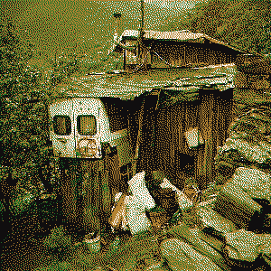 This picture again shows an old hut or this time I would call it a cabin, since it's bigger. It's made of an old bus which has the logo of anarchism written upon it. The cabin is made up totally by used materials mostly wood mixing them together to a pleasent experience of an anti-consumers eye, giving the woo a meaning to live again. Some smoke is coming out of a chimney. The cabin lays in some valley, you can see the ridge line covered with pine trees.
